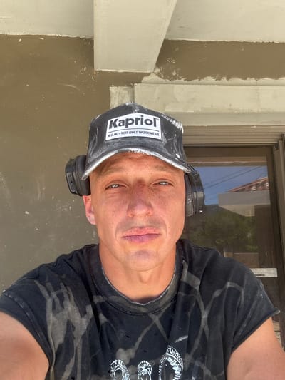 The image shows a close-up, waist-up shot of a person wearing a black cap with white lettering that reads "Kapriol N.I.W. - NOT ONLY WORKWEAR". The person also has black headphones over their ears and is wearing a black t-shirt with a faded, abstract pattern. The background consists of a textured, light brown wall with some white trim and a window or doorframe. The overall impression is of a person engaged in work, possibly outdoors given the lighting and the reflection in the window. The location is specified as Lastva Grbaljska, Montenegro.