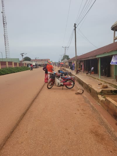 This image depicts a street scene in Tangu, Nigeria. The street is paved with a reddish-brown asphalt material. On the right side of the street, there is a row of buildings with corrugated metal roofs, suggestive of market stalls or shops. A concrete drain runs along the front of these buildings. Several motorcycles are parked along the street. In the foreground, a person wearing an orange shirt and a colorful hat stands next to a motorcycle. Further down the street, other individuals are visible, some near motorcycles and others interacting with the market stalls. On the left side of the street, a tall telecommunications tower is prominent against an overcast sky. There is also a low wall and some vegetation. A light-colored dog is lying on the concrete drain in the middle ground. The overall impression is of a busy, active street in a Nigerian town.