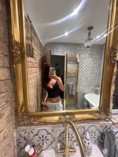 In a bathroom in Brighton, United Kingdom, a person is taking a mirror selfie. The bathroom features exposed brick walls on the left and patterned tiles on the right. A large, ornate gold mirror hangs on the wall, reflecting the scene. The person is wearing a black crop top and white sweatpants. A gold towel rack with towels, a bathtub, and a small chandelier are visible in the reflection. The sink area in the foreground shows a gold faucet, toothbrushes, and various toiletries.