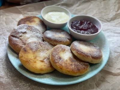 A close-up shot shows a light blue plate filled with seven golden-brown syrniki (cheese pancakes) dusted with powdered sugar. Two small white ceramic bowls are placed on the plate, one containing a creamy white substance, likely sour cream or yogurt, and the other filled with a dark red berry jam or compote. The plate is set on a textured, light brown paper surface, with more powdered sugar scattered around the plate, suggesting it's a freshly prepared dish. The lighting is soft, casting gentle shadows, and the overall impression is one of a homemade, comforting dessert. The scene is likely set in a home kitchen or dining area within Мирновское сельское поселение, Ukraine, given the context of the prompt. There are no people visible in the image, and no specific actions are depicted beyond the presentation of the food.
