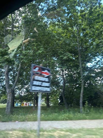 The image depicts a street-level view, likely taken from a moving vehicle, in the city of Beloslav, Bulgaria. In the foreground, a blurred gravel path runs horizontally across the lower portion of the frame. To the left, large, colorful letters spelling "BELOSLAV" are visible, with some letters obscured by trees.
Dominating the mid-ground is a dark signpost with a white rectangular sign attached. The sign features text in what appears to be Cyrillic script, along with a red arrow pointing to the right and a circular symbol with a diagonal line, possibly indicating a prohibition. The sign is somewhat blurred due to motion. Behind the sign, trees with lush green foliage fill the background, with patches of blue sky visible through the canopy. Some of the trees have thick, dark trunks. There is a thin, vertical pole in the center of the image, likely supporting the sign. The overall impression is of a rural or suburban setting with significant tree cover.