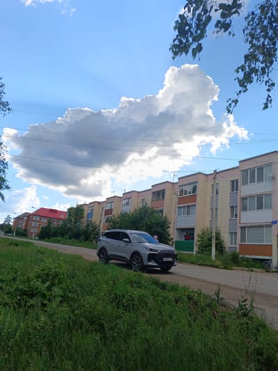 In Tolbazy, Russia, a silver SUV is parked on a dirt road next to a grassy embankment. The SUV faces right, partially obscuring a row of apartment buildings. The buildings are multi-story structures with light-colored facades, some featuring balconies. Green trees are interspersed between the buildings. A large, fluffy white cloud dominates the blue sky. The foreground is filled with lush green grass and some taller weeds. In the background, more apartment buildings and trees are visible, suggesting a residential area within the town.