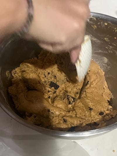 This media file shows a close-up, overhead view of cookie dough being mixed in a stainless steel bowl. A person's hand, with a bracelet visible on the wrist, is seen holding a fluted, white tool, possibly a dough scraper or spatula, and is in the process of mixing the dough. The cookie dough is a light brown color with visible dark chunks, likely chocolate chips or raisins, mixed throughout. The dough appears slightly wet and has a textured, uneven surface. The background is blurred and shows a white surface, suggesting a kitchen counter or table. The lighting is dim, indicating it might be evening or an indoor setting with limited light. There is no visible text in the image. The scene appears to be in a domestic setting, likely a kitchen, and the activity taking place is the preparation of cookie dough. Given the location context of Kalijaten, Indonesia, this is an indoor scene consistent with home baking.