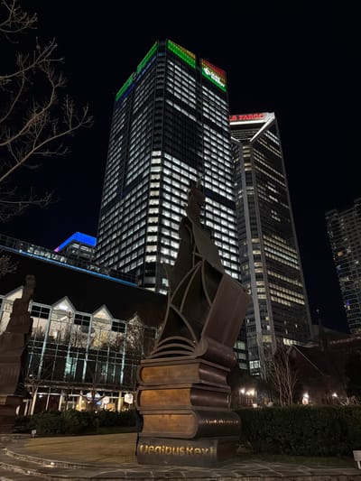 A nighttime view showcases illuminated skyscrapers. One building features a large "UW" design formed by selectively lit windows. Another nearby tower displays "WELLS FARGO" vertically. In the foreground, a bronze sculpture of stacked books is visible, with the title "Oedipus Key" inscribed on one. Bare tree branches are silhouetted against the night sky, and glimpses of a building with sharp roof lines are also seen.