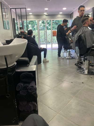 The image displays the interior of a hair salon or barbershop situated in Plaisir, France. The floor consists of light gray square tiles. The ceiling is white with multiple recessed circular lights. At the rear, large glass double doors open to an exterior view of dense green foliage.

In the foreground, a white ceramic hair washing basin sits on a black pedestal. Beneath it, a black storage unit holds several rolled black and dark gray towels. The lower portion of a person's legs, wearing gray pants, is visible at the bottom center, indicating the photographer's seated position.

To the left, a male individual is seated in a black armchair, facing the glass doors. He is dressed in a dark top, dark ripped jeans, and light-colored athletic shoes with white socks.

In the mid-ground, two male barbers are actively engaged with clients. One barber, wearing a dark, paint-splattered long-sleeved shirt, stands while cutting the hair of a client seated in a black barber chair. This client is draped in a black and white striped cape. The second barber, wearing glasses and a dark short-sleeved top, stands to the right, holding a hair clipper. He is positioned near another client, also draped in a black and white striped cape, whose head shows a fade haircut in progress. Additional barber chairs with chrome bases and wall-mounted mirrors are visible within the establishment. A black metal frame structure is present near the glass doors on the left.