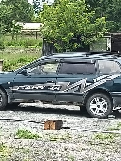The image displays a dark green station wagon parked outdoors on a gravel surface. The car has silver decals on its side that appear to read "CALD JA". A dark brown brick is lying on the gravel in front of the car. The background consists of green trees and a fence. There are no people visible in the image. The scene appears to be in a rural or semi-rural area, possibly a yard or undeveloped land, and the car shows signs of wear and tear. The location in the city of Monastirische, Russia, is not directly evident from the visual information in the image itself.