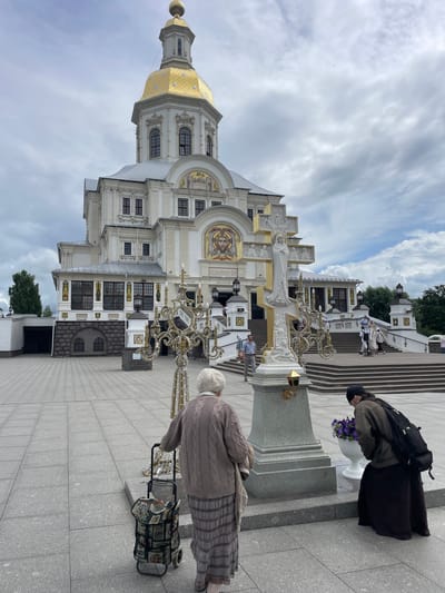 In front of the Holy Trinity Cathedral in Diveyevo, Russia, two women are observed in the foreground. The woman on the left, an elderly individual with white hair, wears a beige knitted sweater and a long striped skirt. She is seen from behind, pushing a patterned rolling shopping cart. To her right, a woman with dark hair and a black beanie bends down, wearing a brown jacket over a long black skirt, and carrying a backpack. She is looking at a white vase filled with purple flowers placed next to a large stone pedestal. This pedestal supports an ornate, gold-trimmed cross with relief sculptures. The cathedral, a grand white structure with a golden dome and mosaics, serves as the backdrop. Several people are visible on the steps leading up to the cathedral entrance. The scene unfolds on a paved plaza under a cloudy sky.