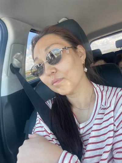 A woman is sitting in the passenger seat of a car, wearing round sunglasses and a red and white striped shirt. Her hair is dark brown and pulled back. She is wearing a pearl earring and a delicate gold necklace. The car appears to be moving, as evidenced by the blurred scenery outside the window and the reflection in her sunglasses, which shows a road and sky. The background suggests a rural or semi-rural landscape, possibly within the Алтынды ауылдық округі, Kazakhstan, given the lighting and vegetation visible. The woman is looking forward, and her expression is neutral. Another person's head is visible in the back seat, and another person's face is slightly visible behind them. A black seatbelt is fastened across the woman's chest.