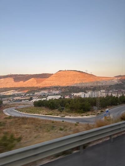 The image captures a wide shot of a landscape featuring a large, terraced construction site on a hillside, likely a quarry or mining operation, in the city of Gülnar, Turkey. The terraced slopes are illuminated by the warm light of what appears to be late afternoon or early morning sun, casting orange and brown hues across the earth.
Below the terraced hill, a settlement is visible, consisting of various low-rise buildings, including residential blocks with multiple stories and industrial-looking structures with flat roofs. Numerous trucks and other vehicles are parked or driving within this area, suggesting ongoing activity. Green trees and dry, scrubby vegetation dot the landscape between the settlement and the hill.
In the foreground, a curved asphalt road is seen winding through the terrain. A silver guardrail runs along the edge of the road. A few cars are visible on the road, with one appearing to be in motion. The sky above is a clear, pale blue, transitioning to a lighter shade near the horizon. Power lines and transmission towers are also present, spanning across the hills and the settlement. The overall impression is of a developing or industrial area situated within a natural, hilly environment.