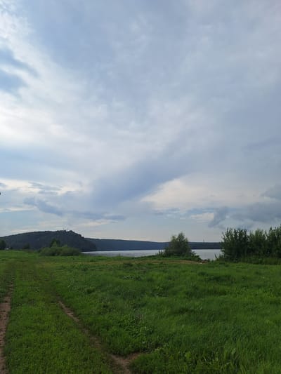 A wide shot shows a grassy field in the foreground, with a dirt path winding through it. In the middle ground, a body of water stretches across the scene, with a forested hill on the far bank. The sky is overcast with dramatic clouds. The scene appears to be in the rural area of Atamanovka, Russia. There are no people or animals visible in the image.