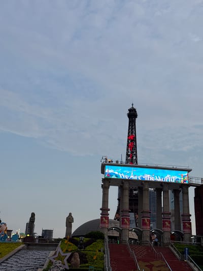The image shows a view of Window of the World in Nanshan District, China. It features a replica of the Eiffel Tower with red Chinese lettering. A large digital screen adjacent to it displays "We Are The World." The surrounding area includes architectural elements, statues, landscaped gardens, and a water feature. The sky is overcast.