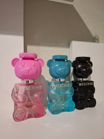 Three Moschino Toy Bear perfume bottles are arranged on a white shelf. From left to right, the bottles are pink, blue, and black. Each bottle is shaped like a teddy bear, with the cap forming the bear's head and the body holding the perfume. The brand name "MOSCHINO" is printed in white on the body of each bottle. The background consists of a textured wall and a white partition. The scene is brightly lit. No people or actions are depicted. The image does not provide any information about the city of Korenovsk, Russia.