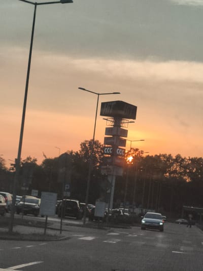 The image shows a street scene at sunset in Nowy Dwór Mazowiecki, Poland. The sky is filled with a warm orange and pink glow, indicating the sun is low on the horizon. In the foreground, there is a road with cars driving and parked. A street lamp is visible on the left side of the image. A multi-tiered sign structure stands prominently in the middle ground, with some of the letters "CCC" visible on one of its panels. Trees line the background, partially obscuring them against the sunset sky. The overall lighting suggests it is either sunrise or sunset.