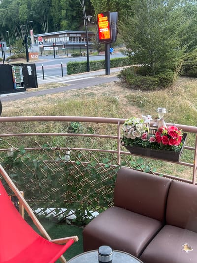 In Maromme, France, a Burger King restaurant is visible with its distinctive orange signage. Adjacent to it, a digital billboard displays an "ATTENTION" warning about a high risk of forest fires, written in French. In the foreground, a brown railing encloses a small patio area. On the railing, a planter box holds artificial flowers, including pink and white roses, and a solar-powered garden light. A red deck chair is positioned to the left, and a brown sofa with visible wear and tear occupies the right side of the frame. A small glass table with a metal object on it is also present. The surrounding environment features dry grass, green bushes, and trees. In the background, a road and a pedestrian crossing are visible, along with a black utility box containing wiring.