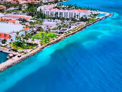 This aerial image captures a vibrant coastal resort scene during the daytime, under bright, clear skies. The dominant feature is the strikingly clear ocean, which transitions from a brilliant turquoise near the shore to a deeper blue further out.
Along the coastline, several large, white multi-story buildings with prominent reddish-orange roofs are visible, appearing to be hotels or resort complexes. These structures are nestled amidst lush green landscaping and numerous tall palm trees, contributing to a tropical aesthetic. Parking lots adjacent to the buildings contain multiple parked cars.
On the right side of the frame, a large hotel complex overlooks a curved, light-colored sandy area or artificial beach that extends into the turquoise water. Further to the left, a small pier or jetty with several covered structures juts out into the water from a rocky stretch of the shore. The coastline in various sections is reinforced with rocky barriers. White lounge chairs or umbrellas are scattered across some of the green lawns, hinting at leisure activities.
The scene suggests a serene and luxurious vacation destination, characterized by its clear waters, sunny weather, and resort amenities. There are no discernible people or specific activities taking place, nor is any visible text present.