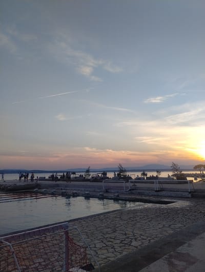 The image depicts a seaside scene in Crikvenica, Croatia, at sunset. A body of water, likely the Adriatic Sea, occupies the lower left portion of the frame, reflecting the sky. The foreground features a paved area constructed from irregular stone slabs, with a white and orange net enclosure, possibly part of a sports field, partially visible in the bottom left corner. Beyond the water, a shoreline is lined with people standing and sitting. A distant, hazy mountain range forms the horizon line. The sky displays a gradient of colors from soft blues and whites at the top to oranges and yellows near the setting sun on the right. A faint contrail from an aircraft is visible in the upper left quadrant of the sky.