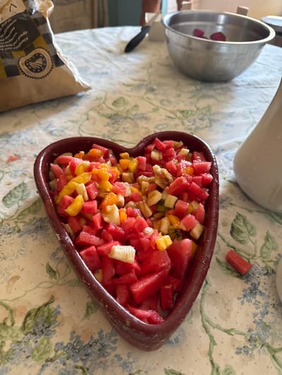 This image shows a heart-shaped dish filled with chopped fruit, including watermelon, kiwi, and possibly banana and orange. The fruit is a colorful mix of reds, pinks, yellows, and greens. The dish is placed on a tablecloth with a floral pattern featuring blue and green flowers. To the left of the dish, there is a metal bowl containing what appear to be slices of strawberry. In the background, a white pitcher or jug is visible. A piece of watermelon is on the tablecloth to the right of the fruit dish. A brown paper bag with some text and a circular seal is in the lower left corner.

The scene appears to be set indoors, possibly in a kitchen or dining area, given the presence of the tablecloth and serving dishes. The lighting suggests it could be daytime. There are no people visible in the image. The overall impression is one of a prepared meal or snack, likely a refreshing fruit salad. The presence of the paper bag with "IGP de..." text suggests the location could be Spain, as indicated by the prompt's location context. The seal on the bag also has text that might be part of a protected designation of origin.