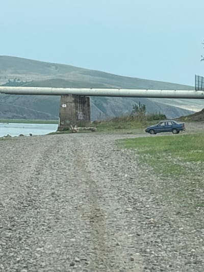 The image depicts a rural landscape scene near Chernogorsk, Russia. In the foreground, a gravel road stretches towards the viewer. In the middle ground, a large pipe, supported by a concrete pillar, traverses the scene horizontally. To the right of the pillar and pipe, a blue sedan is parked on a grassy area. Beyond the pipe and to the left, a body of water, likely a river or lake, is visible, with hilly terrain in the background under a light blue sky. The overall environment suggests an outdoor, undeveloped area.