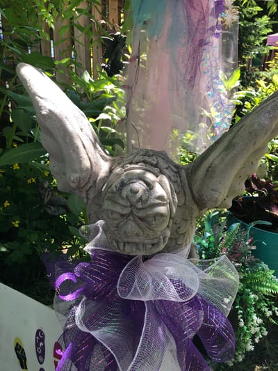 A stone statue of a gargoyle with wings is decorated with purple and white mesh ribbon bows. The gargoyle has glitter on its face and wings. It is surrounded by green foliage, a wooden fence, and some sheer, iridescent fabric. The statue is located outdoors.