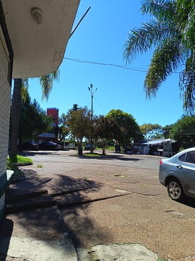 The image shows a parking lot or wide street in Termas del Daymán, Uruguay, on a sunny day. The surface is cracked and worn, made of small pebbles or gravel set in concrete. The sky is a clear, bright blue. Palm trees are visible on the right side of the image, partially framing the scene. A light-colored car is parked on the right, partially cut off in the frame.

To the left is the corner of a building with a white, slightly curved roof overhang and brick exterior. Further in the background, across the street, are more buildings, including a red one with a tower-like structure. There are other cars parked in the distance. Several trees with green foliage are scattered throughout the scene. A lamppost stands in the middle ground.