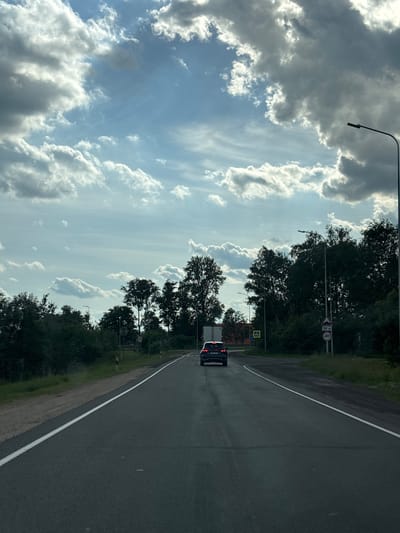 This media file shows a view from a car driving on a paved road, likely in the Лугаваслабадскі сельскі Савет area of Belarus. The road curves to the right, with trees and greenery lining the left side. The sky is partly cloudy with dramatic cumulus clouds scattered across a bright blue expanse. Sunlight filters through the clouds, suggesting it is daytime, possibly late afternoon.
A dark-colored vehicle, possibly a van or SUV, is visible ahead on the road. Traffic signs are present along the roadside. One sign is circular with a red border and a number 40 inside, indicating a speed limit of 40. Another sign is a yellow diamond shape with a black symbol, possibly a warning sign. A lamppost is also visible on the left side of the road. The overall scene appears calm and ordinary, depicting a drive through a rural or semi-rural setting. There is no indication of any specific event or emotional tone, beyond the peacefulness of a scenic drive.