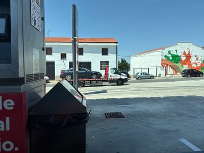 The image shows a street scene in Berrioplano / Berriobeiti, Spain. In the foreground, a red and black garbage bin is visible. Behind it, a tow truck is transporting a dark-colored car. Further in the background, a white building with an orange tiled roof stands, and another white building with a colorful mural of green and orange paint splatters is on the right side of the image. Several cars are parked or driving on the street. The sky is clear and blue, indicating a sunny day.