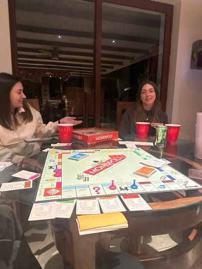 A group of people plays Monopoly around a round glass table. A Monopoly board sits prominently on the table, surrounded by property cards and money. Red plastic cups and a green Monster Energy drink can are placed next to the board. Two smiling women face the camera from opposite sides of the table. The game appears to be in progress, with player pieces scattered across the board. A window is in the background.