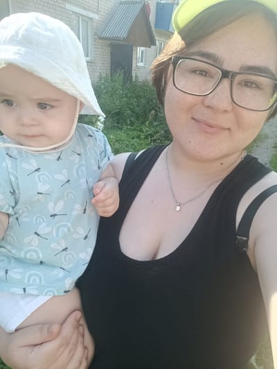 A young woman wearing a black tank top and glasses is holding a baby. The baby is wearing a white sun hat and a light blue t-shirt with dragonfly and rainbow patterns. They are outdoors, in what appears to be a residential area. In the background, a brick building with a metal-roofed extension is visible, along with some greenery and tall grass. The lighting suggests it is daytime. The image was likely taken in the city of Посад, Russia.