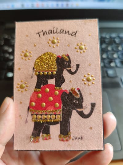 The image shows a close-up of a rectangular card with a pink textured background. The card features a drawing of two elephants stacked on top of each other. The elephants are black with gold and red decorative elements, including glitter on their headdresses and trappings. The top elephant wears a gold glittered headdress and has a red blanket with gold dots draped over it. The bottom elephant also has red and gold trappings and is adorned with gold dots and glitter. The card has the word "Thailand" written in black cursive script at the top, and the word "Jaab" written in black cursive script near the bottom right corner. Several small gold dots and clusters of gold dots are scattered across the card. The card is held by a person's hands, with the fingers of the left hand visible on the left side and the thumb and fingers of the right hand visible on the bottom right. The background appears to be a dark surface, possibly a laptop keyboard, with blurred lights visible. There is no information in the image to suggest the location in the city of Rybnitsa, Moldova.