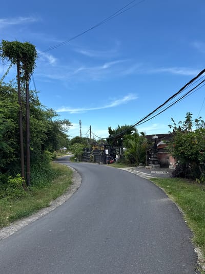 The image depicts a narrow asphalt road curving through a landscape in the city of Kutuh, Indonesia. The sky is a clear blue with faint wispy clouds. Power lines stretch across the upper portion of the frame. On the right side of the road, lush greenery and a traditional Balinese building are visible. The road surface appears well-maintained. On the left, overgrown foliage covers utility poles, adding to the naturalistic aesthetic. The perspective suggests the photo was taken at ground level. There are no visible people or other subjects clearly identifiable in the scene.