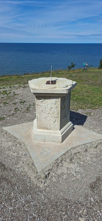 A sundial monument stands on a star-shaped base on a pebbly ground. The sundial face is engraved with Roman numerals and has a gnomon. The stone pedestal of the sundial is decorated with reliefs. In the background, the blue sea meets the horizon under a blue sky with scattered clouds. Two people are visible in the distance, standing on a grassy slope near the sea. The location is identified as Panga, Estonia.