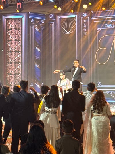 A stage setting features a backdrop with ornate, geometric patterns and visible stage lights. On stage, a man in a black suit with a white stripe on the lapel gestures, while a woman in a floral shirt stands beside him. A crowd of people with their backs turned to the camera faces the stage. Many are dressed in formal attire, including suits and embellished gowns. The angle and perspective suggest the photo was taken from within the audience.