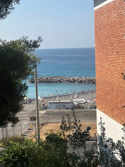 The image depicts a sunlit view of the Ligurian Sea coastline in Ospedaletti, Italy, as seen from a slightly elevated vantage point. In the foreground, dark green foliage from pine and other trees frames the left side of the scene. To the right, a section of a red brick building with a white lower section dominates the frame.
Beyond the foliage and building, a sandy beach is visible, populated with several individuals enjoying the day. Some are seated under umbrellas, while others are in the water. Beach amenities, including small structures and beach umbrellas, are scattered across the sand. A breakwater made of large rocks extends into the turquoise water, protecting a section of the bay. The sea itself is a clear blue, meeting a pale blue sky with no clouds.
In the middle ground, a paved area separates the beach from a fenced-off area with construction fencing and some dry, sandy ground visible behind it. A solitary motorcycle with a black top case is parked on the paved area. A concrete pole with overhead wires is positioned near the center of the image, partially obscuring the view of the beach and sea. The overall impression is one of a coastal town on a clear, warm day.
