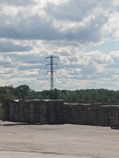 In the city of Okean, Russia, a wide shot captures a large outdoor storage area. In the foreground, stacks of wooden pallets are neatly arranged, stretching horizontally across the frame. Beyond the pallets, a dense line of green trees forms a natural boundary. Dominating the background is a towering electrical pylon, intricately laced with a network of power lines that crisscross the sky. The sky above is a dramatic canvas of billowing white clouds against a pale blue backdrop. The overall scene is one of industrial utility set against a natural landscape under a vast sky.