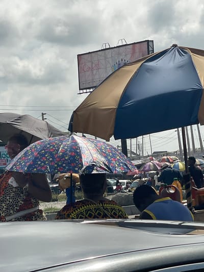 This image captures a vibrant, rotated street scene, likely a market, in Satellite Town, Nigeria, under an overcast sky. The photograph appears to be taken from inside or near a vehicle, looking out onto a busy thoroughfare.
In the prominent foreground, a large, two-toned umbrella, dark blue on top and tan underneath, dominates the left side, angled upwards. Next to it, an open, floral-patterned umbrella with blue, red, and yellow hues provides shade. Below this umbrella, a person is visible, their head and shoulder obscured by the umbrella, wearing clothing with striking, patterned fabric. Another individual in a bright blue top with yellow trim is seen to the right, facing away, close to the side of a white vehicle that frames the right edge of the photo.
The background is filled with the activity of what appears to be a bustling market. Numerous other umbrellas, some striped and multi-colored, are spread throughout, sheltering vendors and shoppers. People are visible amidst the stalls, engaging in daily market routines. Utility poles and a network of overhead wires are noticeable against the cloudy sky, indicating an urban or semi-urban setting.
High above the market, a rectangular billboard stands out against the clouds. The word "Confetti" is clearly visible in a decorative script font on the billboard. Below the floral umbrella, a small green sign with partially visible text like "REN" and "ON THE" can be glimpsed, along with the number "25" on a smaller white sign. The cloudy conditions suggest daytime, possibly a cool or overcast day, conducive to outdoor market activities.
