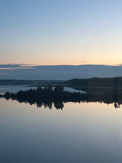 This is an image of a calm lake at dusk, with a small island covered in trees in the middle. The sky is a soft gradient of blue and pink, with a few wispy clouds. The water perfectly reflects the island and the sky, creating a mirror image. The surrounding landscape appears to be hilly and forested. The location is identified as the city of Sudak, Russia, though the specific body of water is not named. There are no people or explicit actions depicted in the image; it's a tranquil, natural scene.
