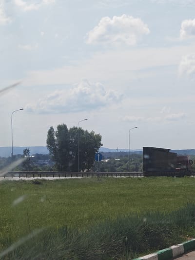 The image depicts a roadside scene on a cloudy day, likely in or near the city of Kurumoch, Russia. In the foreground, a lush green field of tall grass stretches across the frame, with a partially visible white and green painted curb at the bottom right. A metal guardrail runs horizontally across the middle ground, separating the grassy area from a paved road.

On the road, a large, dark-colored truck is moving to the right. It appears to be a tractor-trailer with a covered cargo box. To the left of the truck, a blue circular road sign with a white arrow pointing upwards is visible. Several tall streetlights are positioned along the road.

In the background, rolling hills covered in trees and vegetation are visible, with some buildings and structures suggesting a town or settlement. The sky is filled with scattered white clouds against a light blue backdrop. The overall impression is of a suburban or rural landscape adjacent to a highway. There are no people clearly visible in the image.