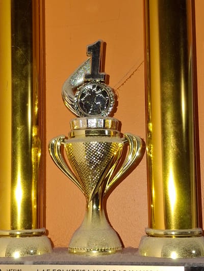 The image shows a close-up of a golden trophy with a silver key attached to it, placed against an orange background. The trophy has a conical shape with gold-colored, curved accents radiating from the top. The base of the trophy appears to be covered in a textured white material. A silver key, with a detailed circular emblem on its head, is positioned horizontally, hooked onto the trophy. The emblem on the key has a black and silver pattern, possibly depicting a crest or a coat of arms.
The overall lighting suggests an indoor setting, with bright reflections on the metallic surfaces of the trophy and key. The background is a plain orange, which might be a wall or a piece of fabric. To the right of the trophy, a white label with black text is partially visible, running vertically. The text is mostly obscured, but the word "LATVIJAS" is legible at the bottom of the label, indicating a Latvian context. The image appears to be a photograph taken in Ogre, Latvia, focusing on the detail and craftsmanship of the trophy and its attached key. There is no clear indication of an activity or event taking place, nor any visible human subjects. The image is likely intended to showcase the trophy as an object of significance.