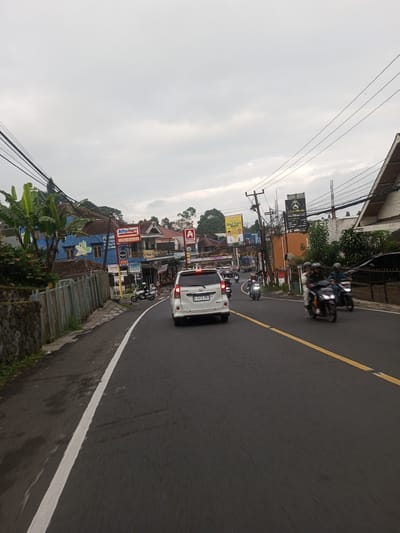 The image depicts a street scene in what appears to be a town or city with medium traffic. A white minivan is driving in the same direction as the viewer on the right side of a two-lane road with a yellow dashed line marking the center. Several motorcycles are also on the road, some alongside the minivan.

Buildings line the sides of the street, with shops and residences visible. Signs for "Alfamart" are prominently displayed on at least two of the buildings, suggesting a chain convenience store. The architecture of the buildings varies, with some having traditional tiled roofs. Power lines crisscross overhead.

The sky is overcast, and the light suggests it might be late afternoon or early morning. Vegetation is visible along the roadside, indicating a semi-urban environment. The exact location within Taunggyi, Myanmar cannot be determined from the visual information provided.