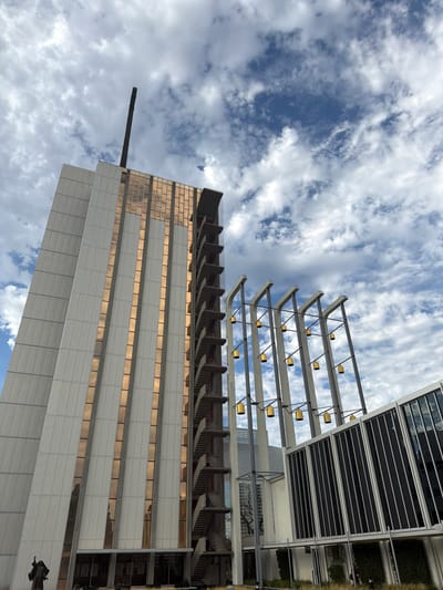 The image shows a low-angle view of the Christ Cathedral in Garden Grove, California, United States of America. The cathedral's tall, white, modernistic spire dominates the left side of the frame, with a dark, slender antenna extending from its peak into the cloudy sky. Vertical strips of gold-tinted glass accent the spire's facade, reflecting the sky. An external concrete staircase with metal railings wraps around the right side of the spire. To the right of the spire, a series of tall, gray concrete columns support horizontal beams from which multiple golden, bell-shaped fixtures hang. Adjacent to these columns, a modern building with a dark, glass-paneled facade is visible. The sky is filled with a mix of white and gray clouds against a blue background. In the foreground, the base of the spire is partially visible, with a dark statue of a robed figure with an outstretched arm to the left. The ground is covered with dry grass.