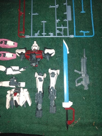 A collection of plastic model kit components is displayed on a dark green textured surface. In the upper-central area, a partially assembled mecha torso with a white head, red chest, and dark grey internal framework is visible. To the upper-left of the torso, three detached armor panels, primarily pink with white accents, are arranged. Below the torso, a white and dark grey backpack or wing assembly is positioned, adjacent to two partially assembled white and dark grey leg units. To the right of the leg units, a sword with a translucent blue blade, white guard, and red grip is laid out. Further to the right, a dark grey rifle weapon is present. In the background, several plastic sprues (runners) are visible: a red one on the upper-left, a grey one centrally, and a partial light blue one on the upper-right. All components are static on the surface.