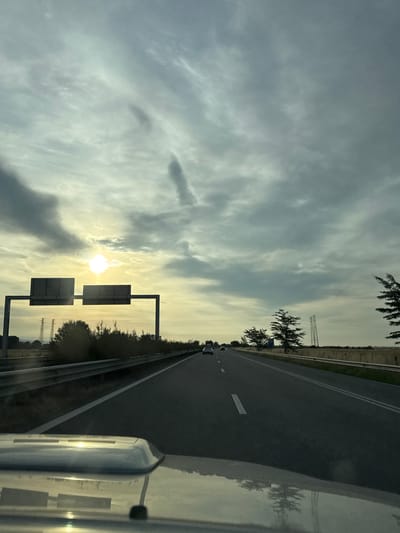 The image captures a view from inside a vehicle traveling on a highway, presumably near Belozem, Bulgaria. The sky is overcast with dramatic, wispy clouds, and the sun is visible as a bright orb low in the sky, casting a yellowish-white light. Two large, rectangular road signs are mounted on a gantry over the highway. The highway itself is asphalt with white lane markings. On the sides of the highway, there are guardrails, vegetation, and in the distance, power transmission towers are visible. The hood of the vehicle is in the foreground, reflecting the sky and the surrounding environment. The overall impression is of a highway journey during daylight with an atmospheric sky.