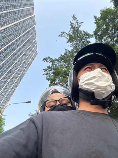 In Bencongan Indah, Indonesia, two individuals are captured in a selfie while riding a motorcycle. The person in the foreground, wearing a black helmet and a white face mask, looks upwards. Behind them, a second person, also wearing a helmet, a black face mask, and glasses, is visible. They are positioned against a backdrop of a tall, modern building with numerous windows and lush green trees. The sky above is clear and blue. The image perspective is from the perspective of someone on the back of the motorcycle.