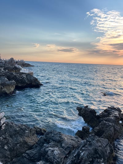 The image shows a rocky coastline on a clear day with a partly cloudy sky. The ocean is a deep blue with small waves lapping against the shore. In the distance, there is a walkway with a railing, and what appears to be beach umbrellas. The sky is a gradient of blue to orange, indicating either sunrise or sunset. The location is identified as Pečurice, Montenegro.