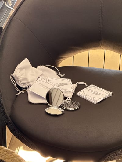 The image depicts a close-up view of a dark upholstered chair, likely a lounge chair, with a textured surface. On the seat of the chair, several items are arranged: two small, white drawstring pouches made of a soft material, possibly velvet, and a stack of three small cards with geometric patterns and text that reads "Live your life, change with courage, yet be yourself!". A silver compact mirror, open and reflecting the surroundings, is placed in front of the cards and pouches. The background shows a hint of vertical wooden slats, suggesting an indoor setting with some decorative elements. The lighting is warm and appears to come from the right, casting shadows on the chair. The image is devoid of any explicit clues or contextual information that would allow for the identification of the city of Sunzha, Russia, or any specific location within it.