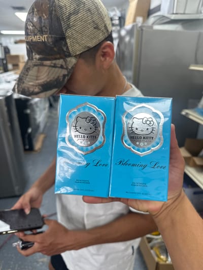A person is holding two boxes of Hello Kitty Blooming Love eau de toilette in what appears to be an indoor, possibly retail, environment. The person is wearing a camouflage baseball cap and a white shirt, and their hands are visible holding a mobile phone along with the boxes. The boxes are light blue with silver Hello Kitty designs. The background includes shelves with various items. The location cannot be determined, and neither can the city Tampa or the United States of America.