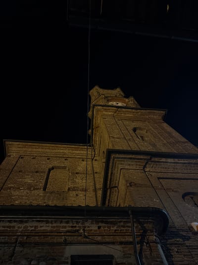 This image captures a low-angle, night-time view of the bell tower of the Church of San Bartolomeo Apostolo in Lagnasco, Italy. The tower is constructed of brick and features a clock face visible towards its apex. Electrical wires are visible running across the dark sky and against the side of the tower. The lower portion of the image shows a section of the church's brick facade with various pipes and electrical conduits attached. The scene is illuminated by artificial lighting, highlighting the textures of the aged brickwork.