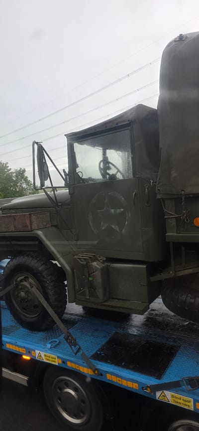 The image shows a military truck, painted olive drab, loaded onto a blue flatbed trailer. The truck has a canvas top over the rear cargo area and a closed cab with a windshield covered in raindrops. A white star emblem is painted on the side of the cab. There is a jerry can mounted on the side of the truck, and a wooden crate is visible on the left side, labeled "FIELD RATION D". The trailer has reflective yellow markers along its sides and straps securing the truck. The ground appears wet, suggesting it has been raining. High-voltage power lines are visible in the background against an overcast sky. The location is identified as Uttlesford, United Kingdom.