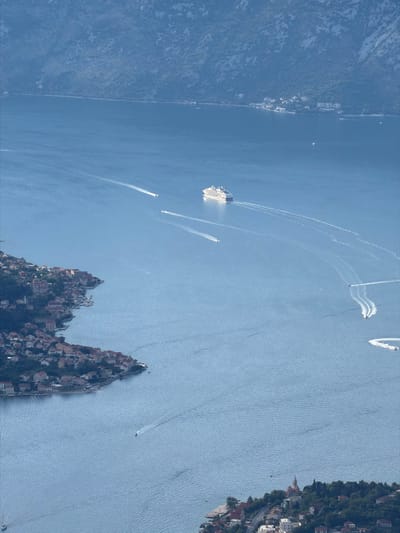 The aerial view captures a vast expanse of deep blue water, likely the Adriatic Sea, with a rugged, mountainous coastline dominating the upper portion of the frame. The mountains are densely covered with vegetation, appearing in various shades of green and brown, with rocky outcrops visible. A cluster of buildings, indicative of a settlement, is nestled along the curved shoreline in the lower left quadrant of the image. These structures are characterized by red-tiled roofs and are situated amidst lush greenery. Several smaller boats are scattered across the water, each leaving a distinct white wake that stretches behind them. In the middle distance, a larger white cruise ship is prominently featured, also generating a significant wake. The water surface exhibits subtle ripples, suggesting a gentle current or breeze. In the lower right section of the image, more buildings are visible, including what appears to be a church with a distinctive dome, further outlining the presence of human habitation along the coast. The overall scene is one of a serene coastal landscape with maritime activity.