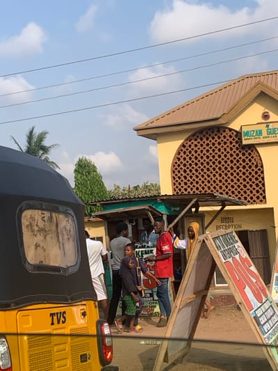 The image depicts a daytime street scene in Nagazi, Nigeria, specifically near Muzan Guest House on Nagazi-Ovete, Adavi L.G.A. In the immediate foreground, the rear and left side of a yellow TVS brand tricycle with a black canopy is visible.

In the mid-ground, a two-story yellow building with a brown corrugated metal roof is prominent on the right, identified by a sign as "MUZAN GUEST, NAGAZI-OVETE, ADAVI L.G.A." This building features an arched window with an intricate dark brown lattice screen on its upper floor, and a "RECEPTION" sign below. To the left of this guest house, a series of single-story commercial stalls with green corrugated metal roofs are operational.

Approximately seven to nine individuals are present in the scene, primarily gathered around the stalls. On the far left, partially obscured by the tricycle, a male in a white t-shirt and white shorts walks away. A group of people stand in front of the stalls, including a man in a grey t-shirt, two children (one in a dark hijab), a young man in a red jersey holding an object and looking left, a man in a light blue shirt behind him, and a woman in a yellow head covering gesturing to the right.

Two A-frame commercial signs are positioned on the unpaved ground to the right of the stalls; the closer sign advertises "KIN KIKENE ENTERPRISES" and "POS" services. Overhead, multiple power lines stretch across a clear blue sky with scattered white clouds. Palm trees and a tall, slender green tree are visible in the background.